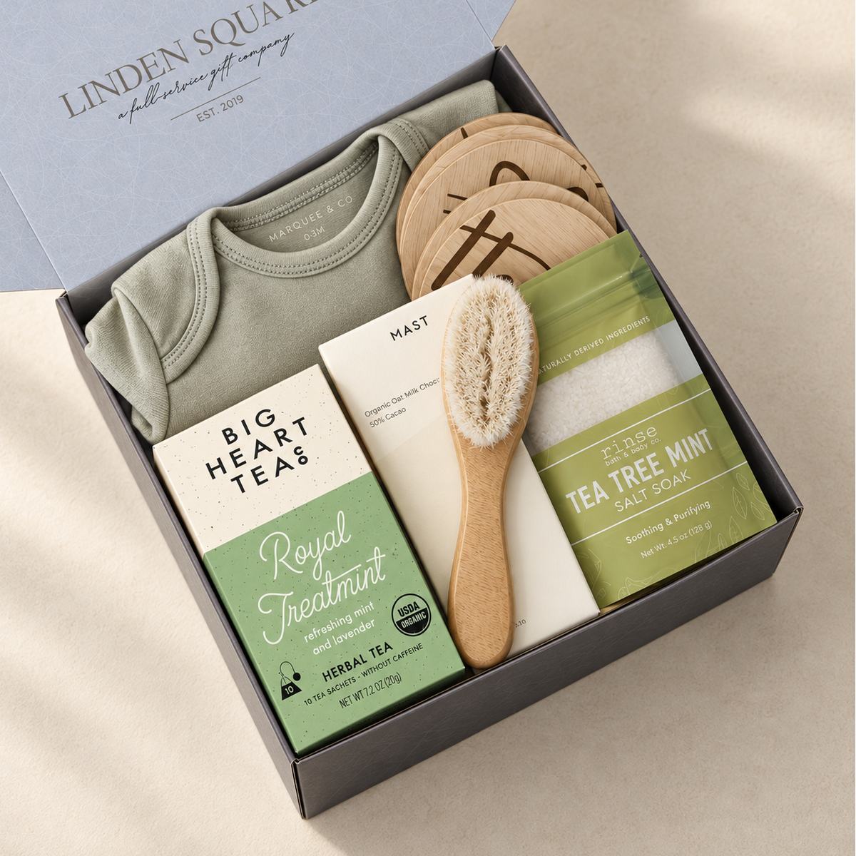A curated gift box set from Linden Square of Allendalept Company, featuring a sage green baby bodysuit, a wooden hair-brush, a box of Big Heart Teas Royal Treatmint herbal tea, and a bar of Nature's Way tea tree mint salt soap, all arranged inside a dark gray box.